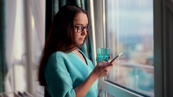 Nervous and Excited Woman is Typing Message By Mobile Phone Standing with Glass Near Window in Room