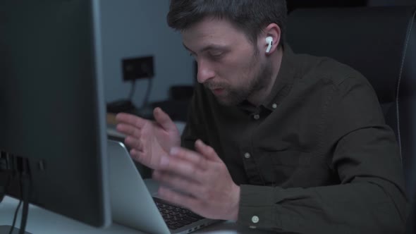 A Male Programmer Works at a Computer and Uses an Inear Headset to Talk with a Colleague or Customer