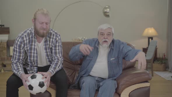 Portrait of Disappointed Caucasian Father and Son Watching Soccer Match Indoors. Adult Man and