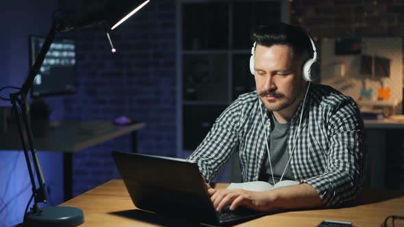 Cheerful Man Enjoying Music in Headphones and Working with Laptop in Dark Office