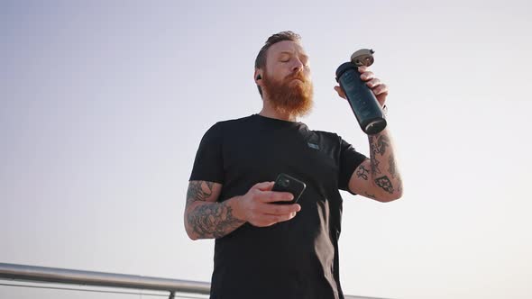 Bearded Man Looks in Smartphone Drinking Water From Thermos