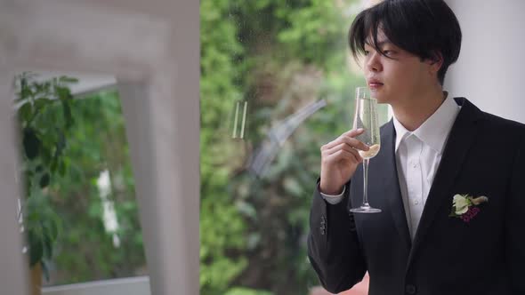 Portrait of Serious Young Asian Man in Wedding Suit Looking Out the Window Drinking Champagne