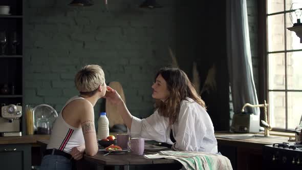 Young Lesbian Couple Is Eating Sandwiches and Kissing at Table in Modern Apartment Spbd.