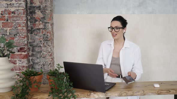 Young Beautiful Woman in Glasses Works at a Laptop and Answers a Phone Call on a Headset