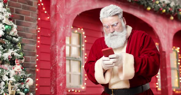 Modern Santa Claus is Playing a Game on a Smartphone on the Background a Red House in a Beautiful