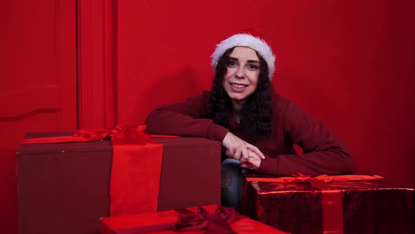 Young Woman in Santa Claus Hat Sitting in Gifts Near Red Wall