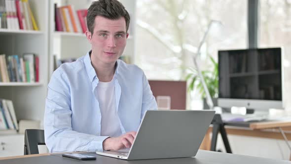 Young Man Smiling at Camera While Working on Laptop