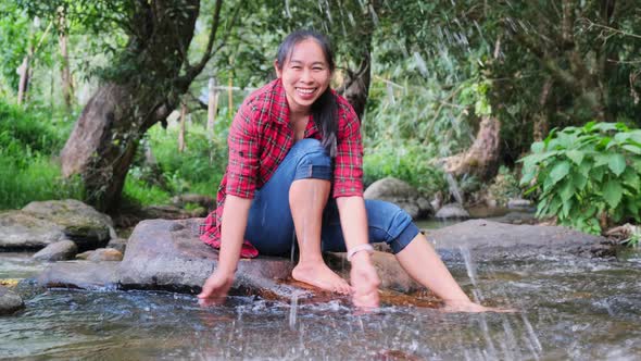 A beautiful Asian woman sits on the stone in the middle of a stream in a mountain forest and playing