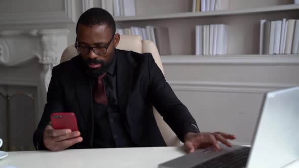 Male Boss Is Sitting in His Working Office, Reading Message in Mobile Phone, Black Man Is Working