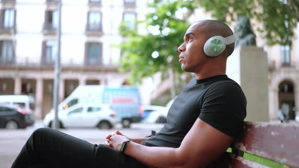 Serious bald African man listening music in headphones while sitting on the bench