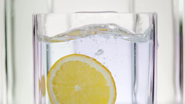 Slice of Lemon Falls Into the Water in a Glass Container on a White Background