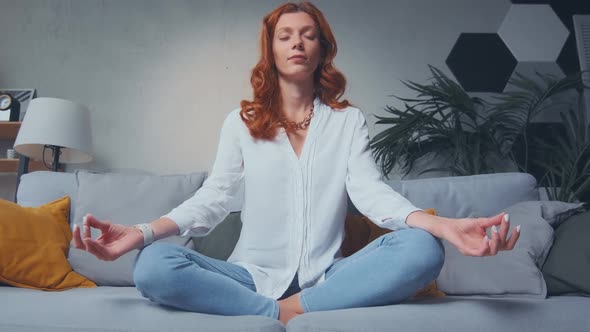 Red Haired Woman Making Mudra Gesture Sitting in Lotus Position on Couch at Home