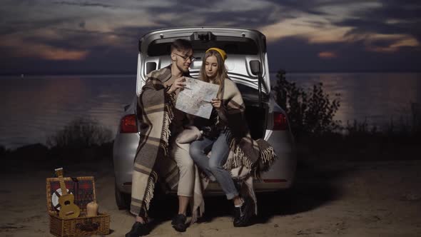 Wide Shot Portrait of Happy Smiling Loving Young Couple Sitting in Car Trunk and Examining Paper Map