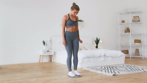 Young Woman in Gray Sportswear Standing on Weight Scale and Rejoicing By Weight Loss Being in White