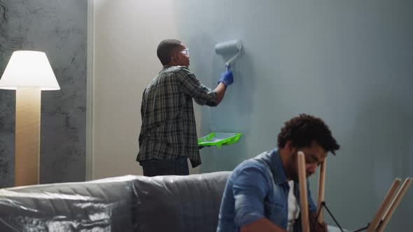 Black Man Applies Paint on Wall Friend Fixes Chair in Room