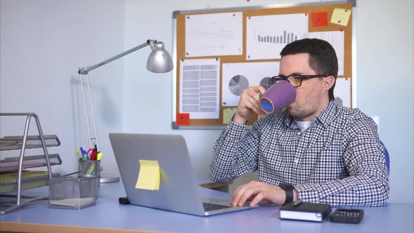 Office Worker Looking at Monitor Drinking Tea While Printing Text on Laptop