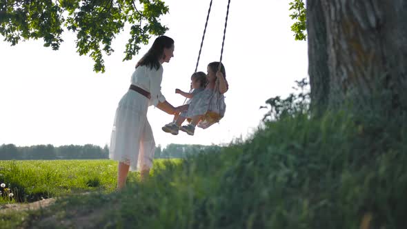 Mom Swings Her Two Happy Daughters on a Rope Swing in the Middle of a Green Field