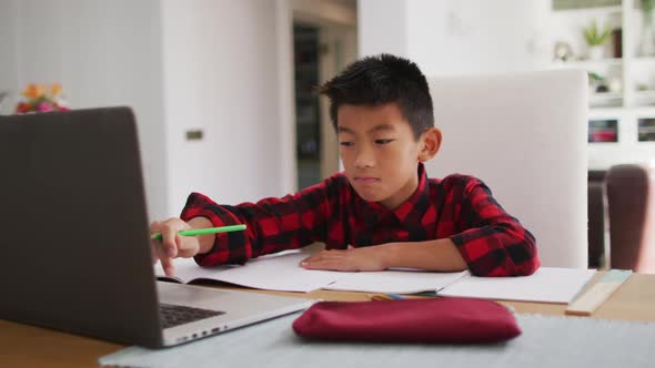 Asian boy at home, sitting at table using laptop for online school lesson
