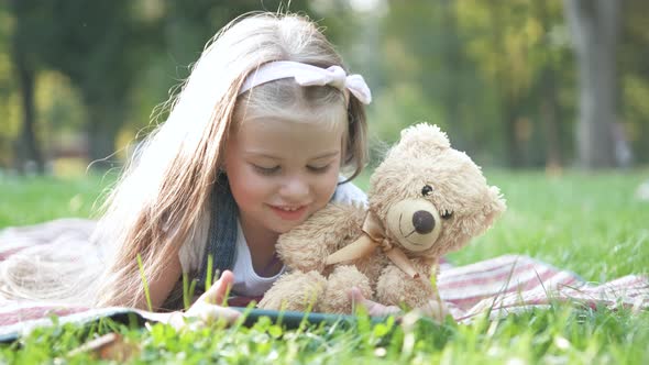 Happy Little Child Girl Playing Her Sellphone Together with Favorite Teddy Bear Toy Outdoors in