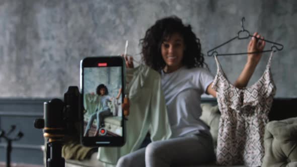 AfricanAmerican Fashion Blogger Recording Video with a Smartphone at Home Choosing Clothes Sitting