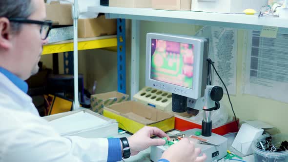 A Man in a White Robe and Glasses Checks the Electrical Board for Defects Through a Magnifying