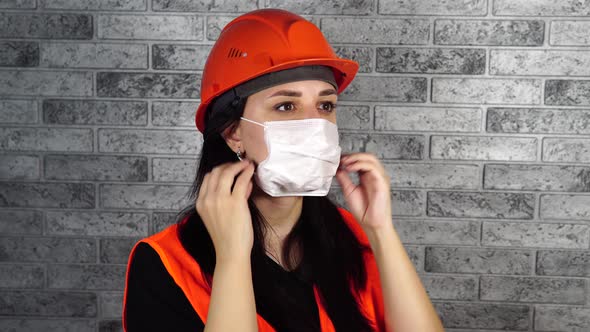 Female Construction Worker in Overalls Putting on Medical Mask on Face on Background of Gray Brick