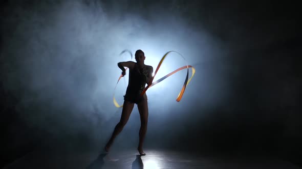 Gymnast with a Ribbon in the Hands Circling in the Smoke. Black Background. Light Rear. Silhouette