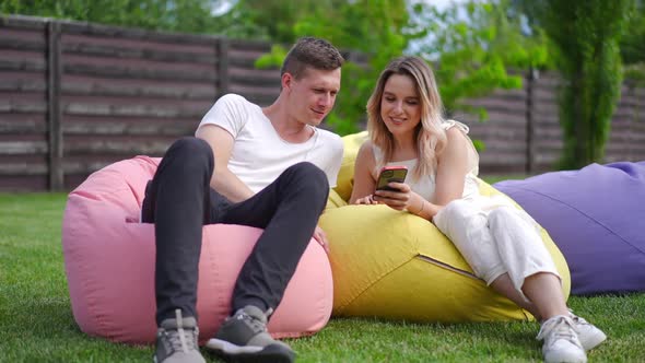 Wide Shot Positive Millennial Couple Sitting on Bag Chairs on Green Meadow Scrolling Social Media on