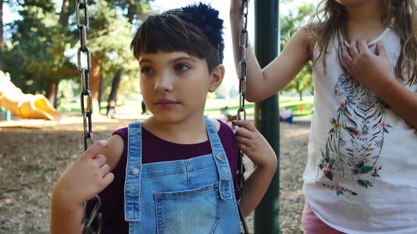 Girl on swing-set shyly looks up as she is bullied by someone out of frame