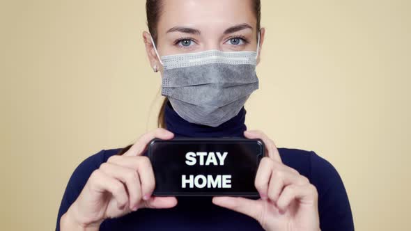 Portrait of Young Woman in Medical Mask Shows Phone with Inscription Stay Home