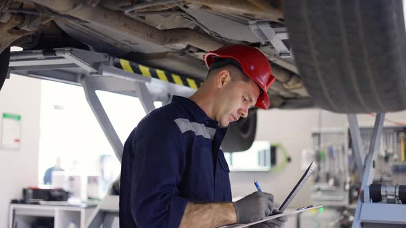 A Car Mechanic Standing Under Lifted Car Making Notes on a Tablet