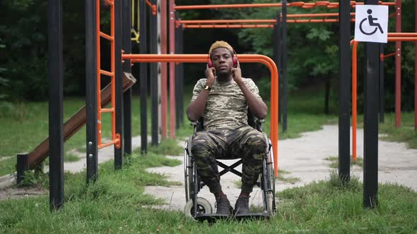 Wide Shot Portrait of Positive Brave Soldier in Wheelchair Listening to Music in Headphones Smiling