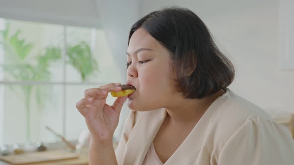 Asian oversize big women enjoy eat unhealthy foods, bite sweet chocolate donut in kitchen at home.