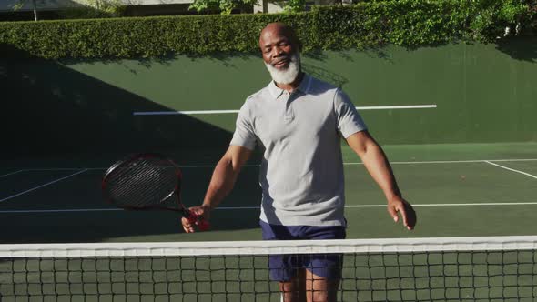 Portrait of african american senior man holding racket smiling while standing on the tennis court