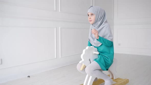 Muslim Teen Girl in Hijab and Dress is Playing Riding on Toy Horse Rocking Chair in Her Room