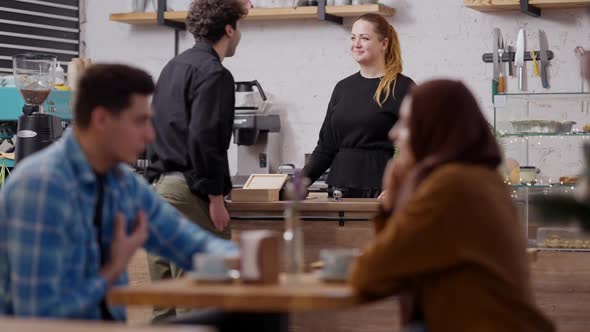 Positive Caucasian Cashier Welcoming Middle Eastern Client in Cafe with Blurred Interracial Couple