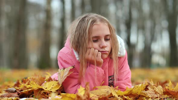 A Little Girl Indifferently Lies on the Autumn Foliage in a City Park