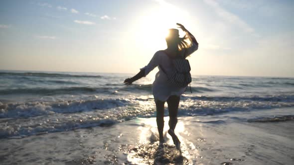 Young Happy Woman in Shirt with Backpack Running To the Sea and Spraying Water with Her Feet at