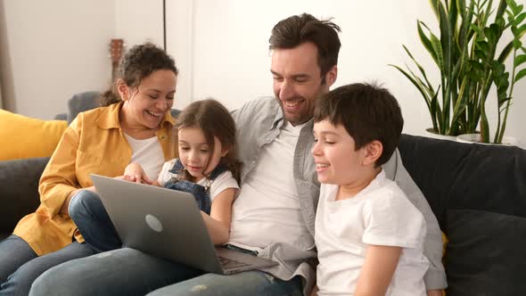 Cheerful Happy Family of Four Using Laptop Sitting on the Sofa in Cozy Apartment