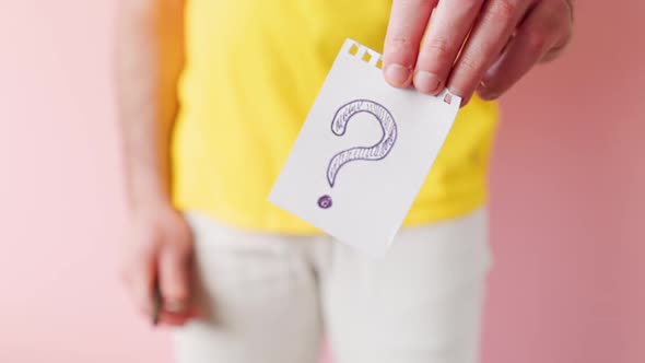 A man in white jeans writes on a piece of paper and shows a question mark drawn on it