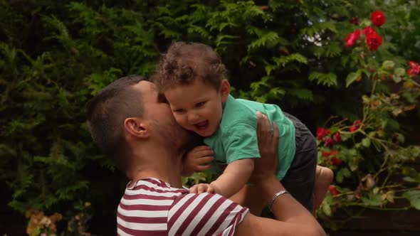 Handsome Dad in a Striped T-shirt Plays with a Curly Baby Boy Outdoors