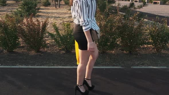 Beautiful girl in black high-heeled shoes, a black skirt and a striped shirt walks through the park