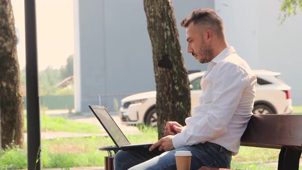 A Business Man in a White Shirt Works on a Laptop While Sitting on a Bench