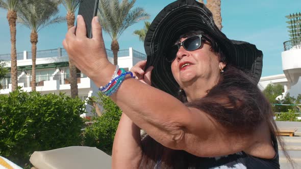 Caucasian Senior Female Elderly in a Black Hat Makes a Selfie on a Smartphone and Sunbathes in the