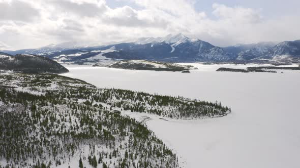 Aerial moving forward to reveal icy, frozen lake surrounded by snow and green pine trees with large