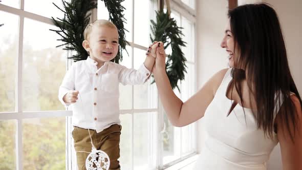 Young Smiling Mother in White Dress is Holding Her Son's Hand While He is Walking on the Window Sill