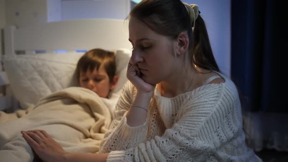 Upset Sad and Stressed Mother Sitting Next to Bed of Her Sleeping Child at Night