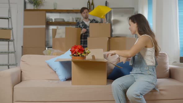 Young Woman Sitting on Couch Packing Belongings in Cardboard Boxes with Boyfriend on Background