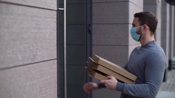 Side view of delivery man wearing medical mask ringing door. Deliveryman holding box with pizza