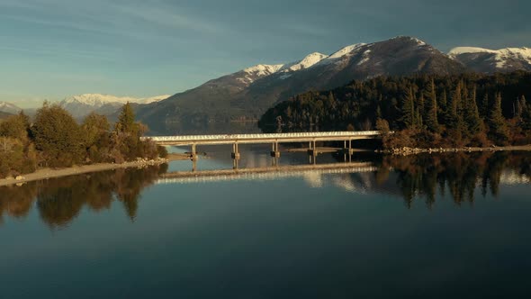 Aerial shot orbiting around a bridge in the middle of a scenic lake with mountains on background nea